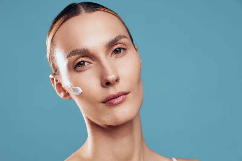 Close up portrait of a calm woman with cream on her cheek, soft lighting, blue background, minimal makeup, highlighting a simple skincare moment
