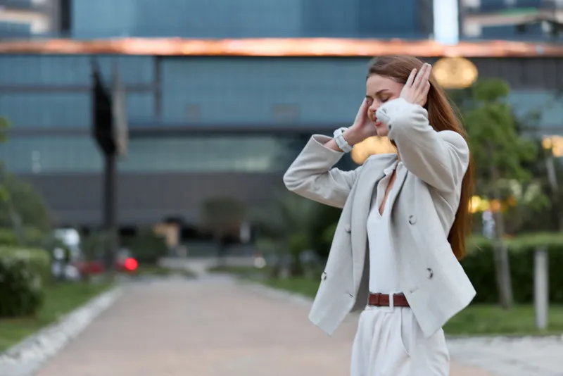 Woman in business attire stressed with hands on head outdoors, expressing psychological pressure and anxiety, urban background, white blazer, fall season