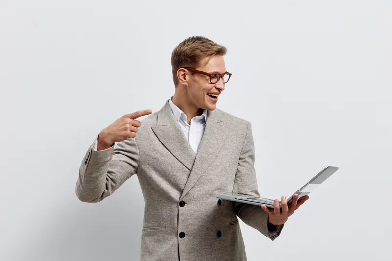 Smiling young businessman with glasses pointing at his head while holding a laptop on a plain white background, expressing excitement or revelation. Studio portrait, professional concept, business suc