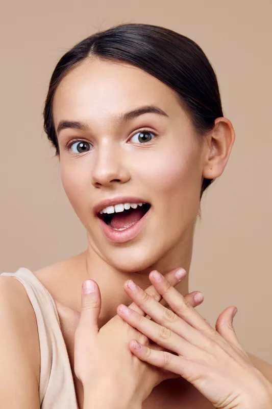 Cheerful young woman with a joyful expression, wearing a light top, showcasing natural beauty against a neutral beige background with soft lighting