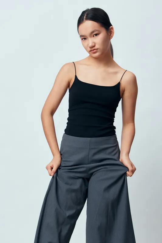 Young woman posing in casual black tank top and wide leg pants, standing with hands in pockets against plain light background for fashion or lifestyle concept.