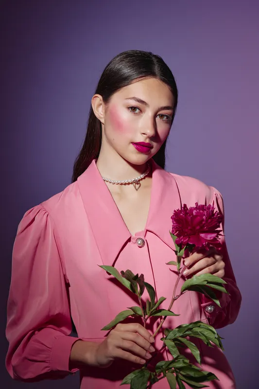 Young woman with long dark hair wears a stylish pink dress and bright red lipstick while holding a vibrant flower on purple background. Beauty creative fashion concept.