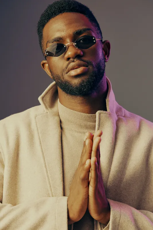 Stylish African male model wearing sunglasses and a beige coat poses confidently with a serious expression, showcasing modern fashion and attitude