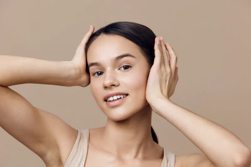 Young woman with natural beauty expressing joy, wearing a beige top, against a neutral background, showcasing healthy skin and a vibrant smile