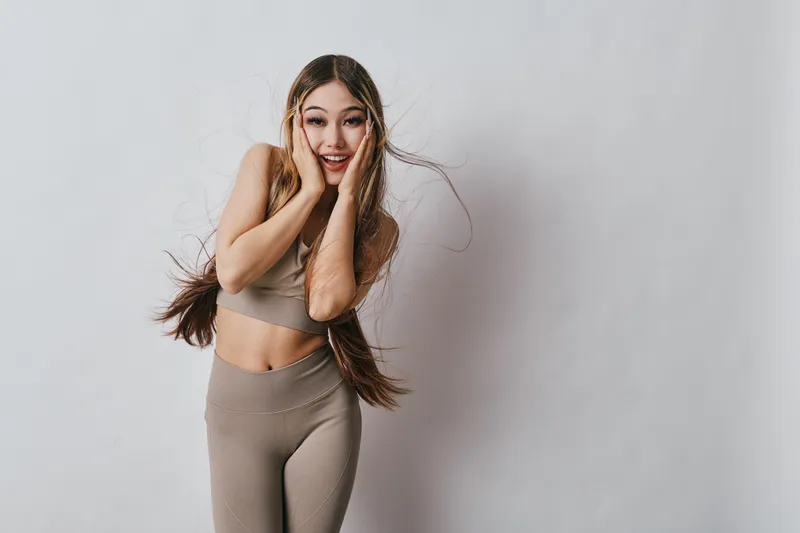 Young woman in beige activewear with long hair posing with surprised expression on white wall background for fitness, lifestyle and casual portrait. Studio shot with natural light.