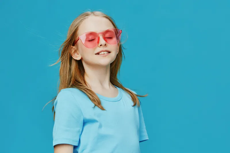 Stylish girl with blonde hair in sunglasses, smiling against a bright blue background, capturing the essence of summer joy and vibrant fashion trends