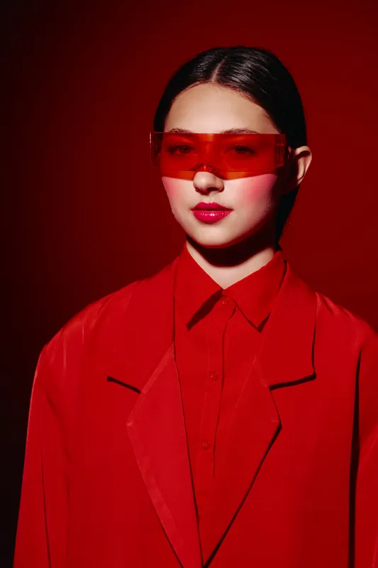 Young woman wearing oversized red sunglasses and a bold red coat poses against a deep red background. Concept of beauty, fashion, and creative portrait.