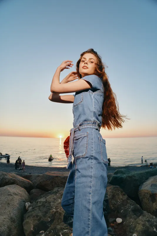 young woman in denim outfit stands on rocky shore during sunset, with long hair flowing and sea horizon in background, casual style and natural light