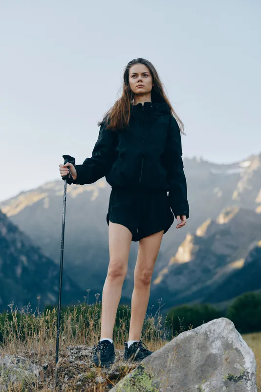 A young woman stands on a rocky trail in a mountainous landscape, gripping a trekking pole. She wears a dark jacket and shorts, ready for an outdoor adventure.