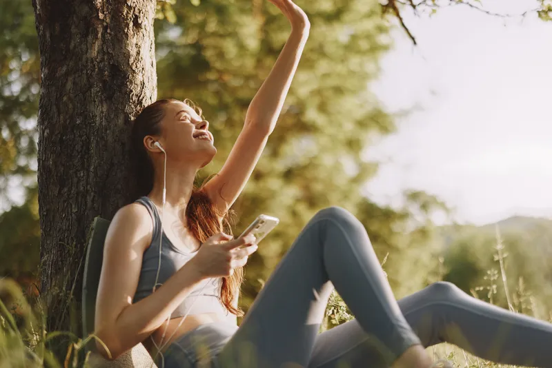 Young woman enjoying music outdoors in activewear, exuding joy while sitting against a tree, with a lush green background and warm sunlight, representing relaxation and wellness
