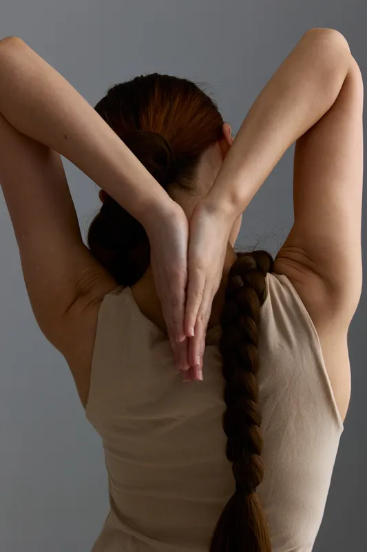 Woman with long braid stretching arms behind head, wearing beige tank top against plain grey background, showing graceful posture and natural skin texture.