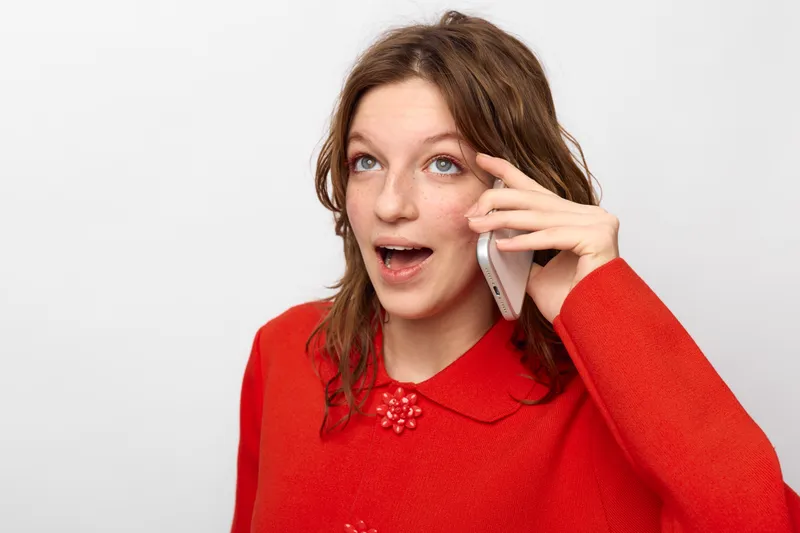 Young woman in red sweater talks on smartphone with surprised expression. Isolated on white background. Concept of communication, technology, emotions, and casual lifestyle.