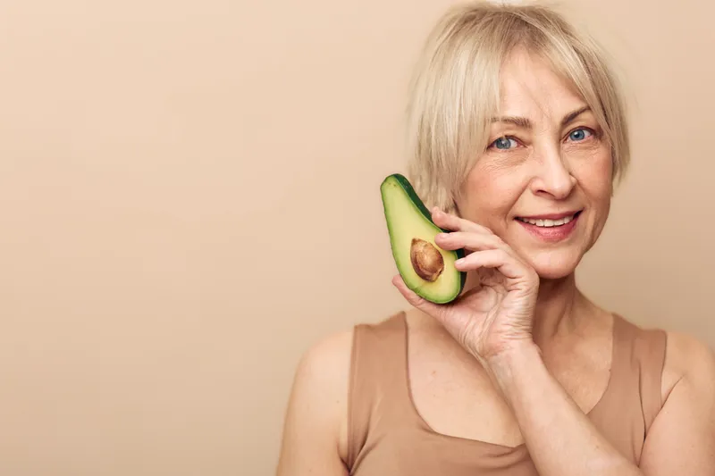 Smiling mature woman with short blonde hair holds fresh avocado slice near face on beige background. Concept of healthy lifestyle, natural beauty, nutrition, and wellness for aging skin.