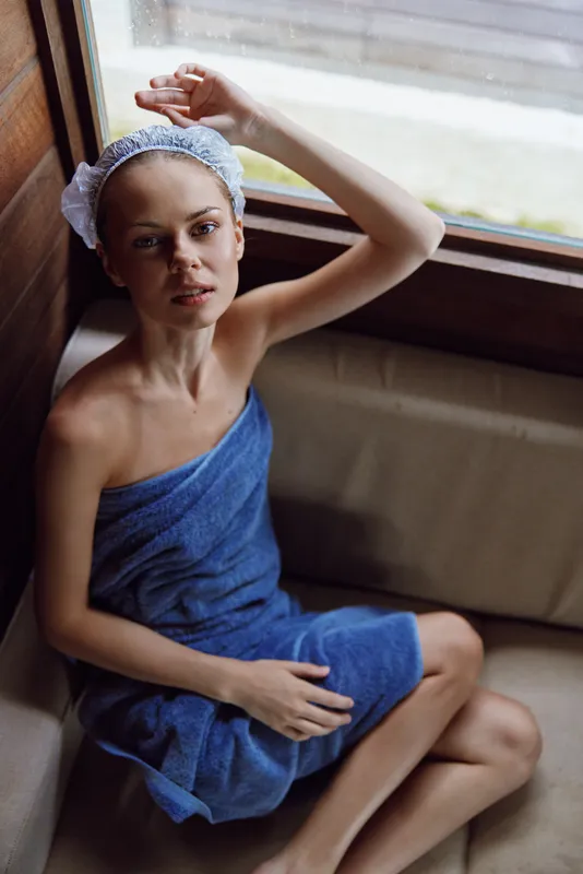 Young woman wrapped in a blue towel with a shower cap, sitting indoors by a window, natural light highlights her relaxed and fresh appearance after bathing.