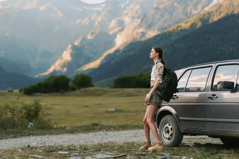 A woman traveler with a backpack stands beside a parked car in a rugged valley, gazing toward distant mountains. An outdoor lifestyle moment of exploration.
