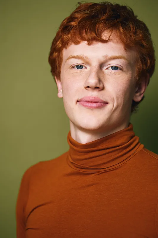 Young man with red hair and fair skin wearing a brown turtleneck sweater posing against olive background with direct eye contact and neutral facial expression.