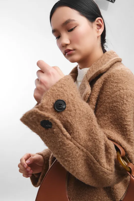 asian woman wearing a tan wool coat adjusts her sleeve in a studio portrait, exuding calm confidence and modern style. The soft lighting highlights textures and creates a poised, professional vibe.