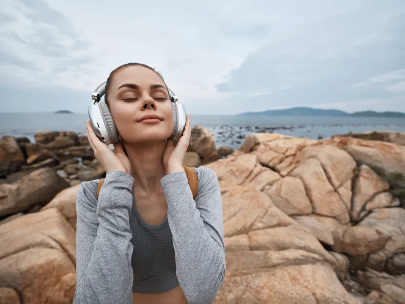 Young woman enjoying music by the ocean, wearing casual sporty clothes and headphones, expressing joy and relaxation amidst a rocky beach background