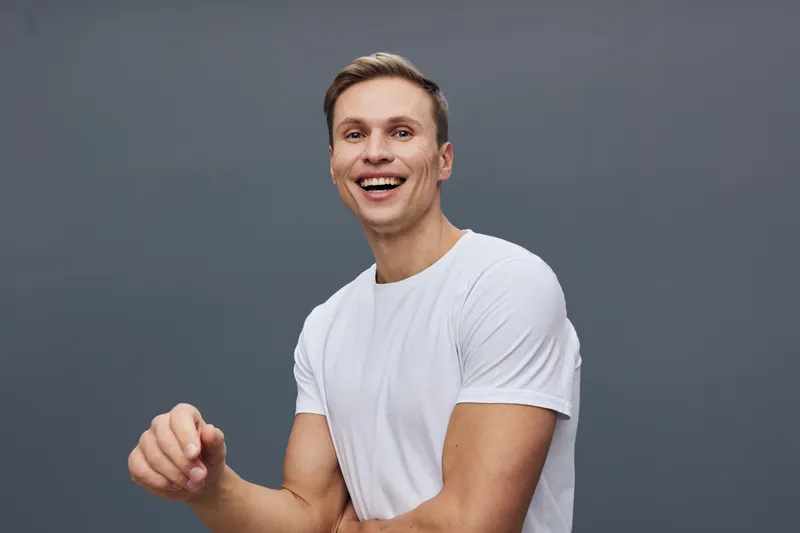 Smiling happy young Caucasian man in white t-shirt looking at camera, expressing joy and confidence, isolated on plain gray background, studio portrait, casual style, positive emotion, lifestyle conce