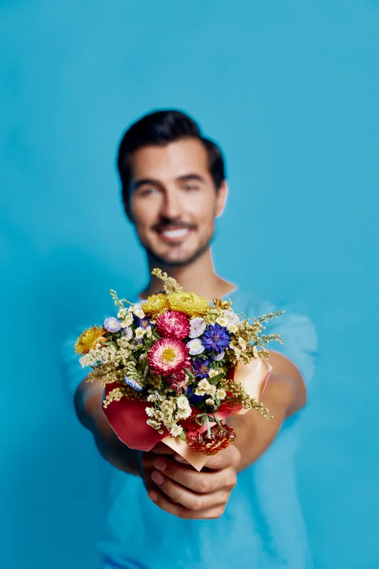 Smiling man holding vibrant flower bouquet in hand, standing in front of bright blue background. Cheerful and positive mood captures heartfelt gesture of giving.