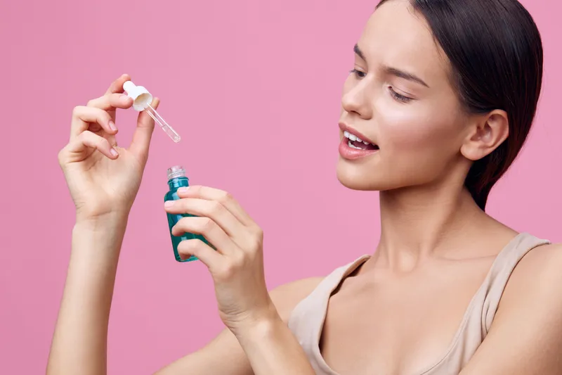 Young woman using a serum dropper over a pink background promoting skincare, beauty, and self care with confidence and joy