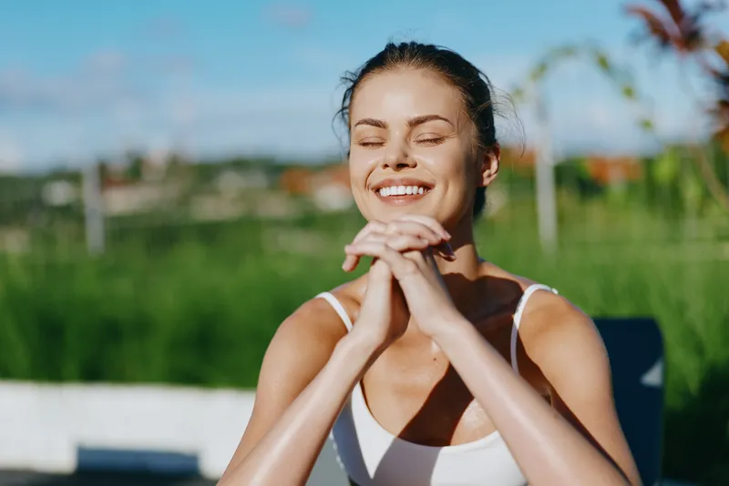 Smiling woman in white tank top enjoys sunny day outdoors with eyes closed and hands clasped under chin, expressing peaceful lifestyle and positive mood in natural setting.