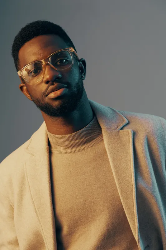 Stylish young Black man with glasses in a cozy beige sweater and light gray coat, expressing confidence against a softly lit blue background