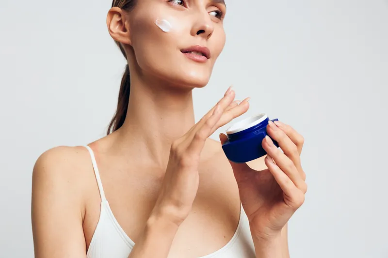 Young woman with long brunette hair applies moisturizing cream on her cheek while holding a blue jar, wearing a white tank top against a plain light background.