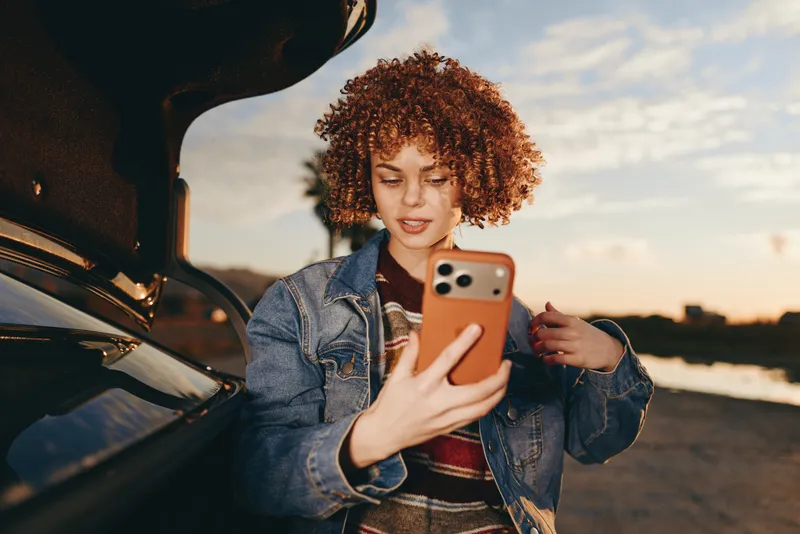 Woman selfie smartphone car sunset curly hair denim outdoor lifestyle, she holds a smartphone by an open car door, warm golden light at the coast, casual travel mood