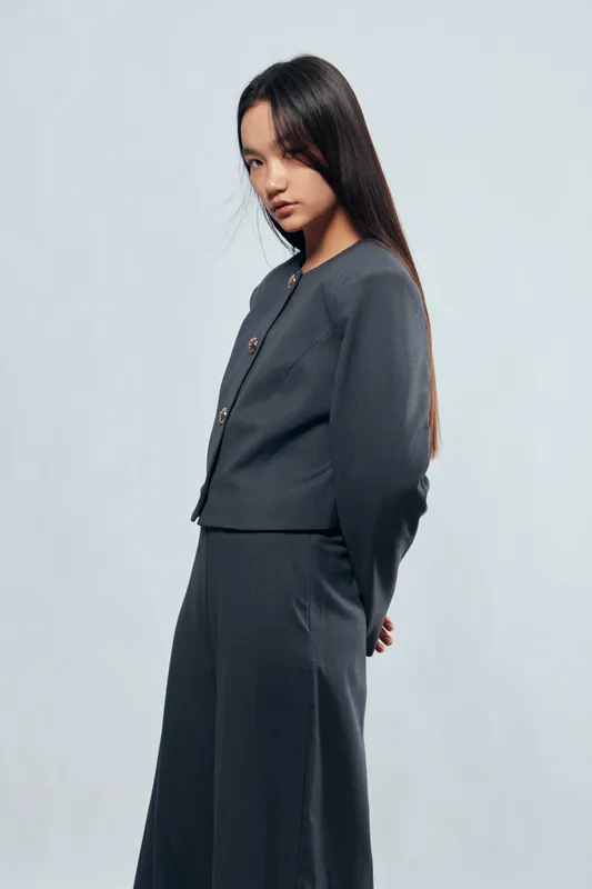 Young woman in dark gray suit standing against light background with hands behind back looking sideways with serious expression and long straight hair in studio portrait.