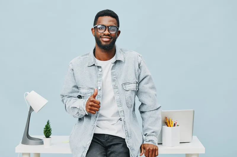 Smiling man with glasses wearing casual denim jacket and white t-shirt standing in modern home office with desk lamp, small plant, and organizer on light blue background. People lifestyle concept