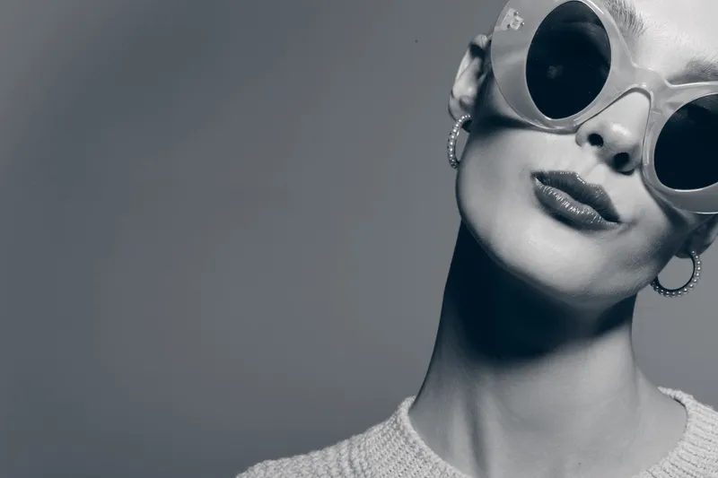 Closeup of a white woman with pale skin wearing round sunglasses and hoop earrings on grey background. Fashion concept with retro style.
