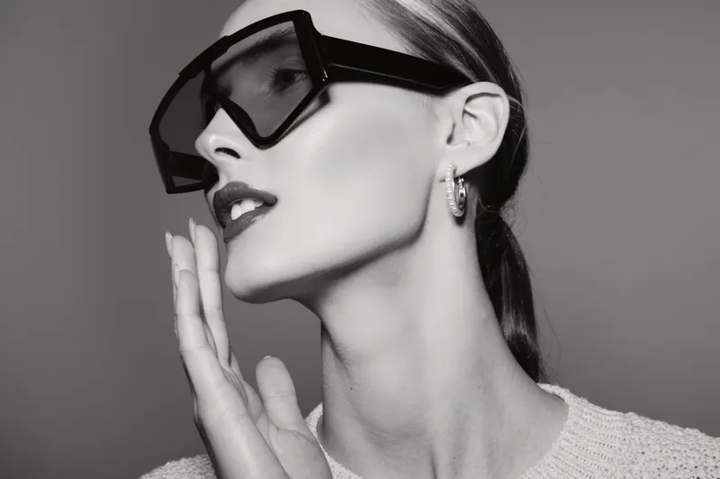 Stylish white woman with light skin wearing oversized black sunglasses and hoop earrings poses in black and white studio portrait with hand near face, fashion concept.