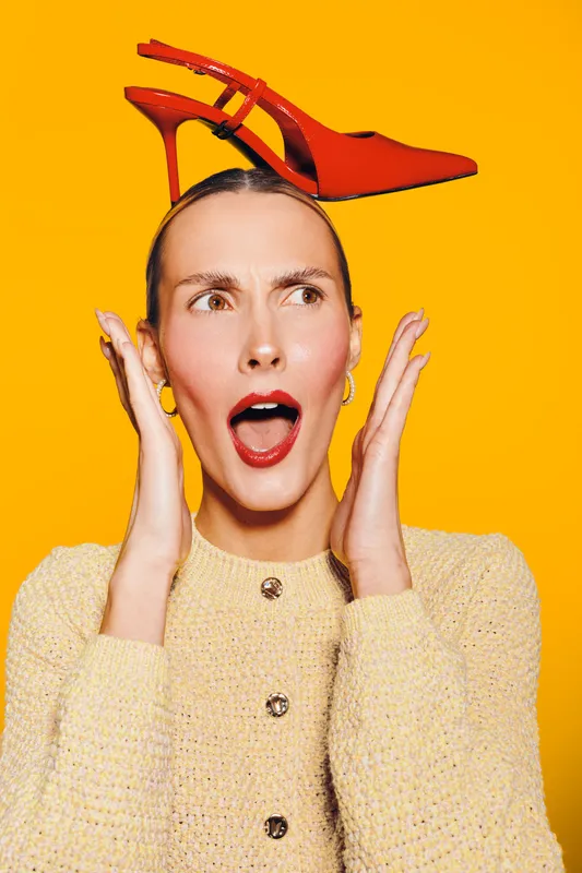 Surprised woman with red high heel shoe on head looks shocked on bright yellow background. Concept for fashion, style, creativity, and unusual accessories.