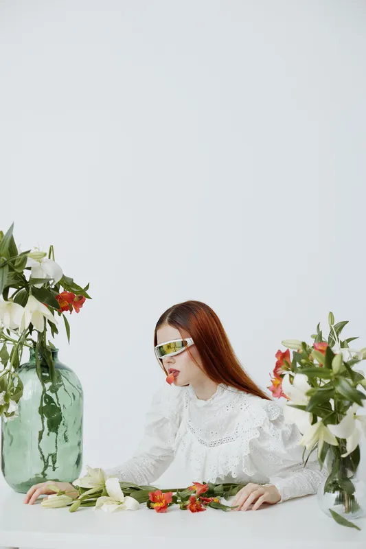 Portrait of a woman with red hair in a white blouse, surrounded by flowers and a green vase, showcasing a futuristic aesthetic and emotional expression