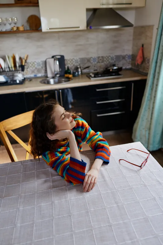 Relaxed woman with curly hair resting her chin on folded arms at a kitchen table, colorful striped sweater, home lifestyle scene, warm natural light, cozy domestic moment.