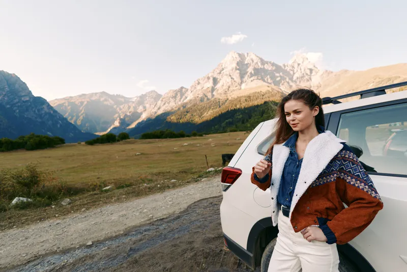 Woman standing by car on mountain road, travel and outdoors lifestyle portrait with scenic landscape. Casual jacket and confident pose near SUV for road trip adventure and exploration.