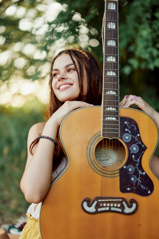 Молодая женщина в стиле хиппи с эко-образом улыбается и смотрит в камеру, держа в руках гитару, на природе во время путешествия