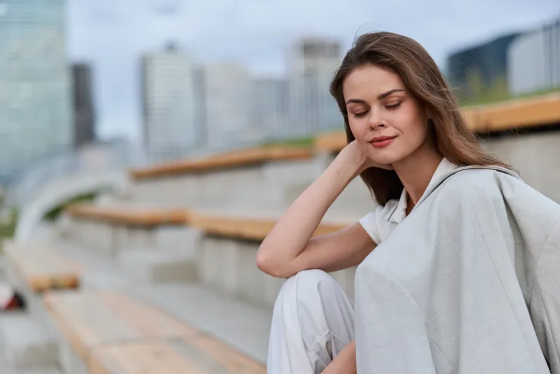 Thoughtful business woman in casual attire sitting outdoors, showcasing modern confidence and professionalism against an urban backdrop.