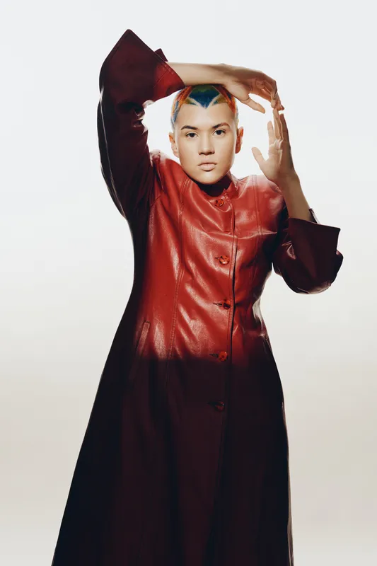 Woman with colorful short hair wearing a shiny red long coat, posing confidently against a plain light background, showcasing modern fashion style and attitude.