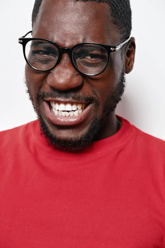 African man with dark skin wearing glasses and red shirt smiling broadly showing teeth on white background. Portrait, happy expression and casual style with closeup face.