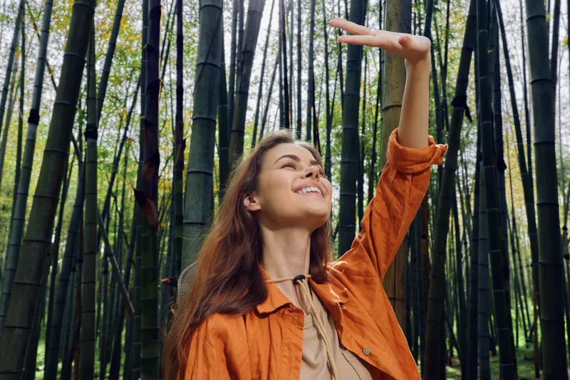 Woman smiling portrait in bamboo forest outdoors, enjoying nature and sunlight while exploring travel lifestyle, casual jacket and relaxed happy expression among tall green trees.
