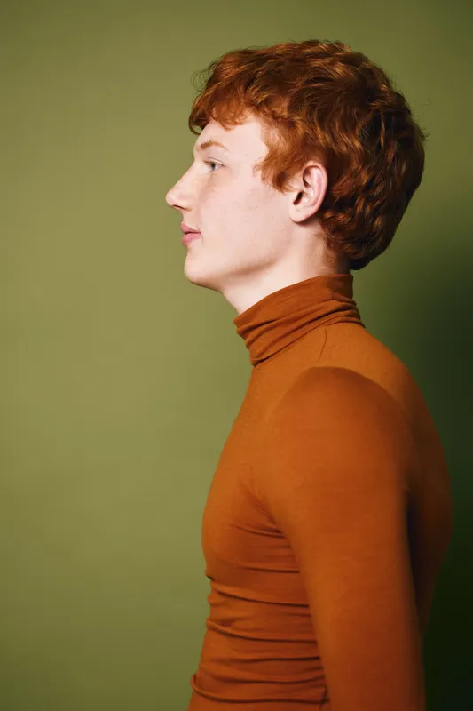 Profile portrait of young redhead man wearing brown turtleneck looking forward with neutral expression on green background in studio lighting with side view.