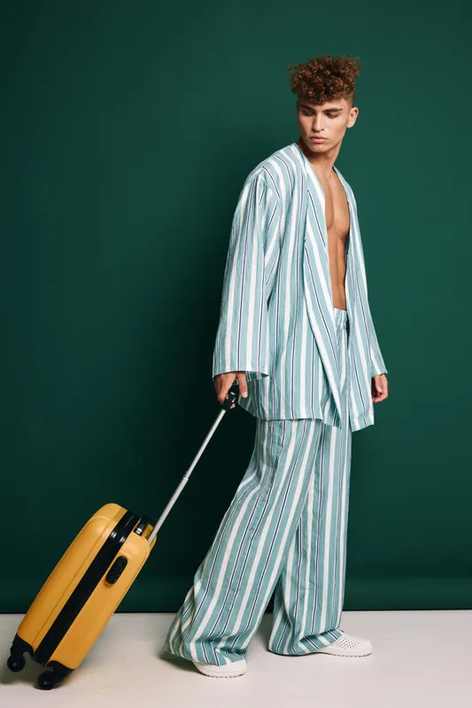 Young man in blue striped oversized suit, casual open wear, dragging yellow rolling suitcase for travel or trip at studio with dark teal background and white sneakers on floor.