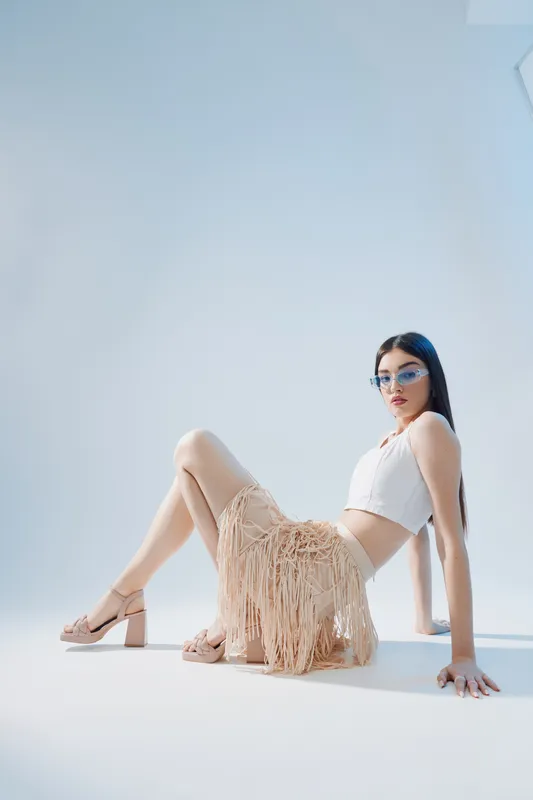 Young woman in fashionable beige fringe skirt and white crop top poses on floor against light background. Concept of beauty, fashion, style, and modern trends in studio.