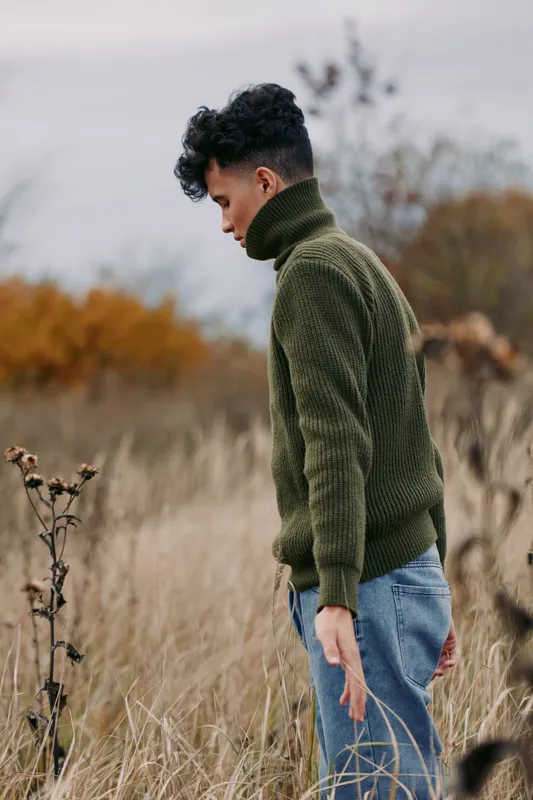 Portrait of a young man standing in a windswept field wearing a textured green sweater, captured as an authentic, natural moment that emphasizes quiet introspection, tactile detail and muted tones.
