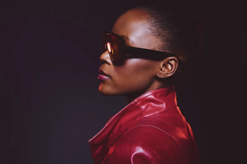 Profile of a confident dark skinned African woman wearing large sunglasses and a red leather jacket against a dark background. Concept of fashion, style, and elegance in portrait photography.