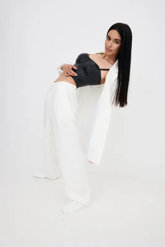 Stylish woman with long black hair wearing white wide-leg pants and a black top stands with a dynamic pose against a plain white background, modern fashion concept.