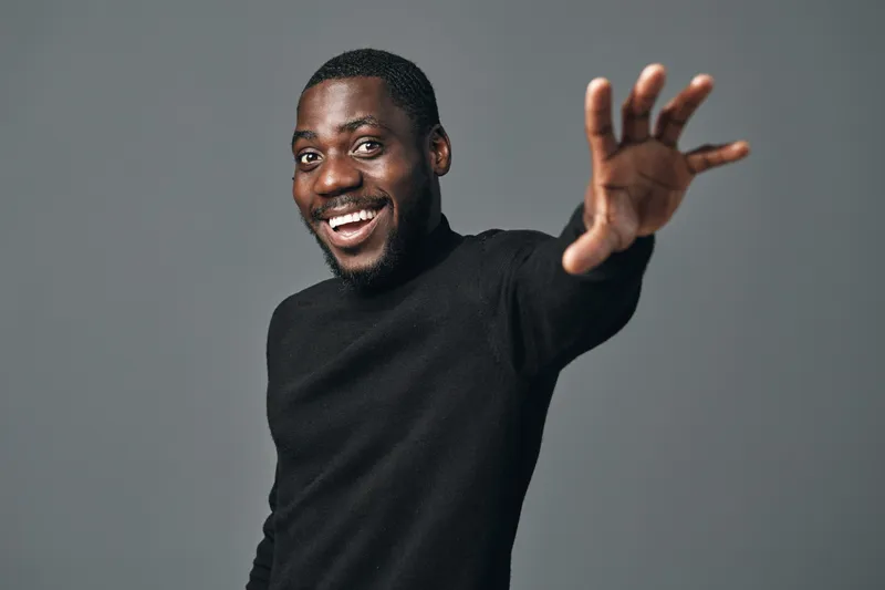 African man smiling and reaching out with hand in creative conceptual visual style. Portrait of happy black male with beard wearing black sweater on gray background.