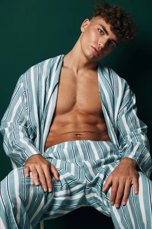 Young man with curly hair wearing striped open shirt and pants sitting with relaxed pose against dark background in studio portrait fashion photography.